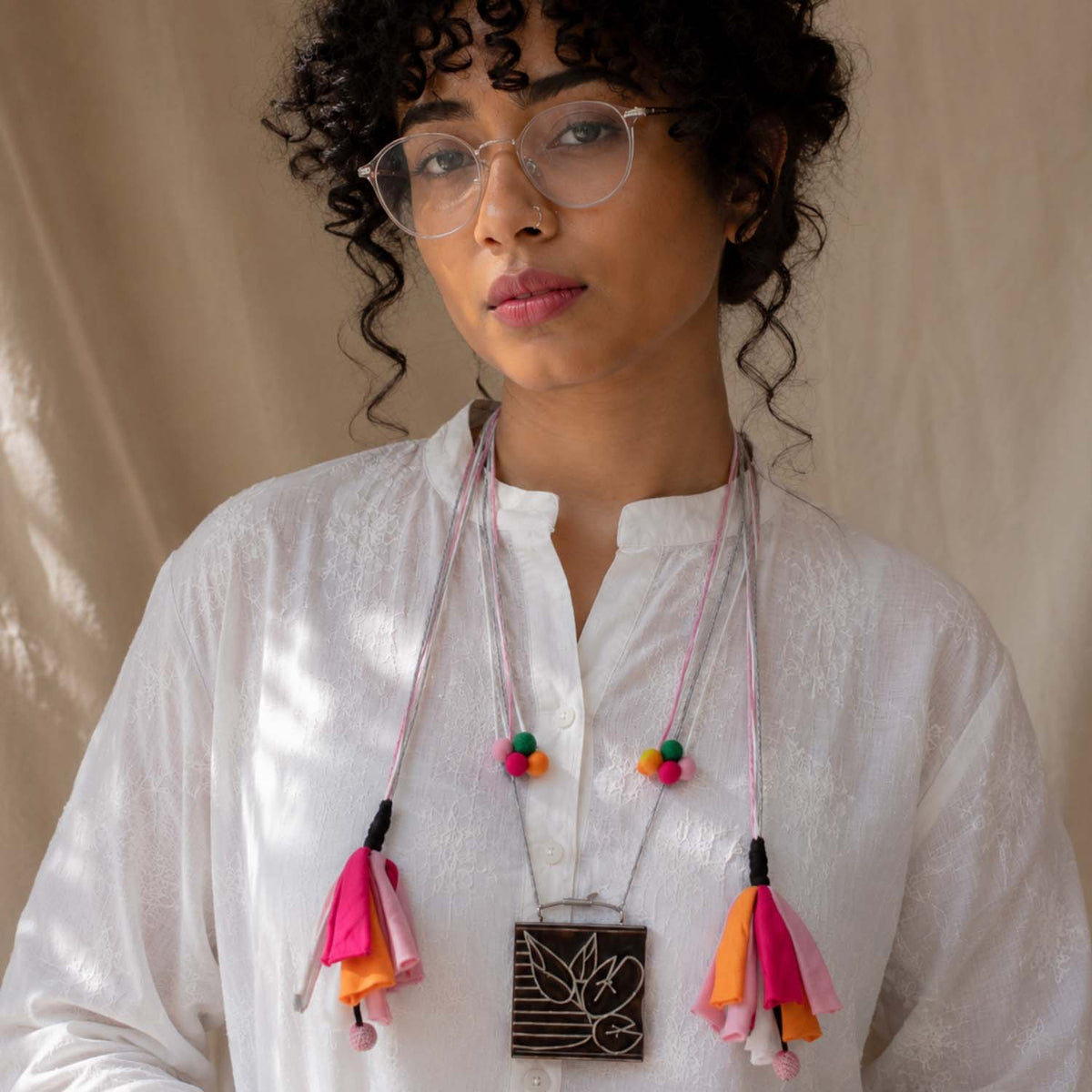A woman with curly dark hair and glasses wearing a white  kurta featuring a long fabric necklace with colorful tassels and a square hand carved wooden pendant.