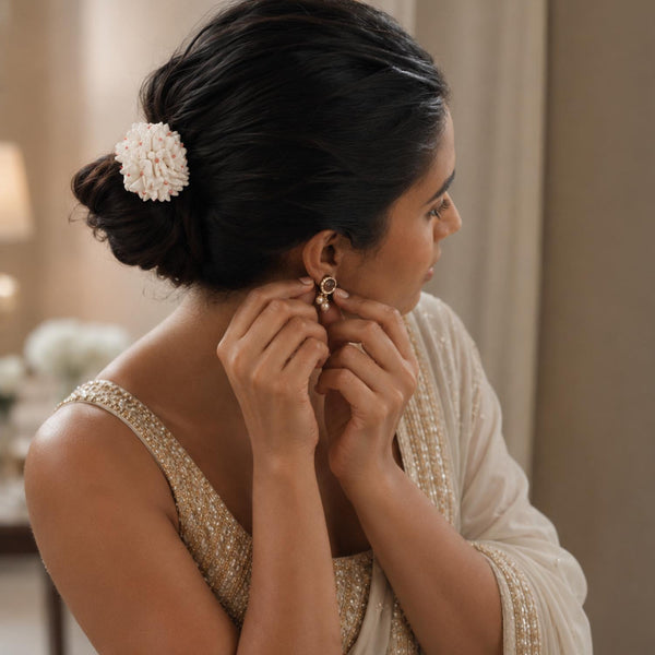A woman with her hair in a low bun and a white shiuli hair accessory is shown from the side and back, wearing a white and gold embellished traditional outfit while adjusting a gold hoop earring.