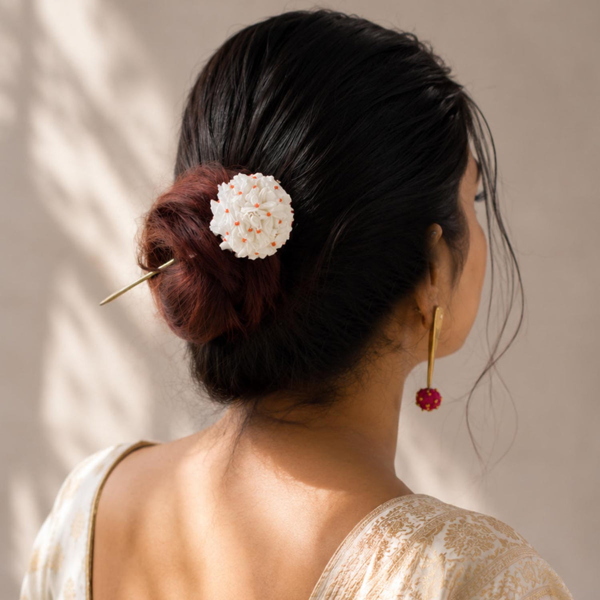 A woman shown from the back wearing a dark hair bun adorned with a White shiuli juda pin hair stick accessory, paired with a red beaded drop earring against a warm beige background with soft leaf shadows.