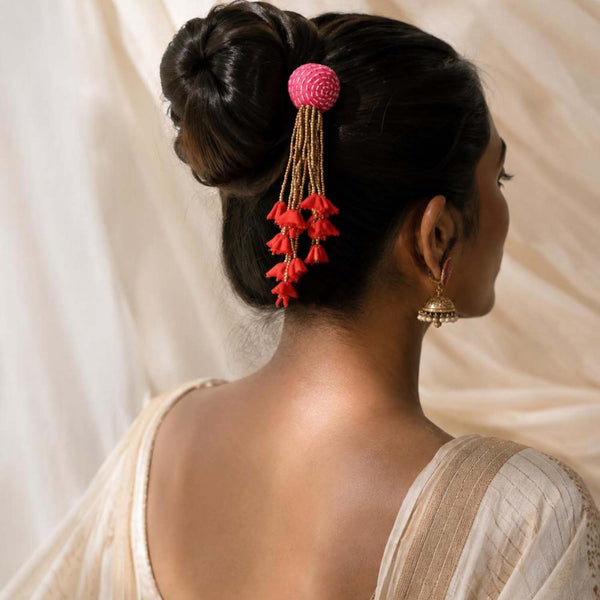 A woman shown from the back wearing a dangling zubeida juda pin,hair accessory with a pink round top, gold-toned hanging strands, and red tiny florals in her dark hair. She is wearing a light beige garment against a soft cream fabric backdrop.