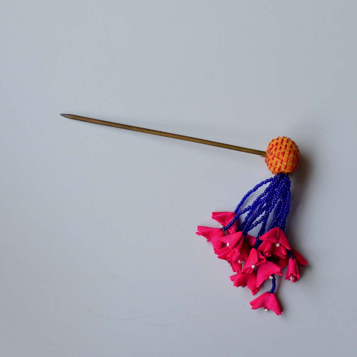 Flat lay of the zubeida dangling juda pin hair accessory that consists of yellow handembroidered fabric bead, blue beads tassles that end in tiny fabric hot pink flowers.
