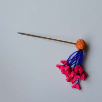 Flat lay of the zubeida dangling juda pin hair accessory that consists of yellow handembroidered fabric bead, blue beads tassles that end in tiny fabric hot pink flowers.