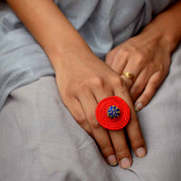 A person's hand wearing a red and blue Rangoli finger ring with a brass band.